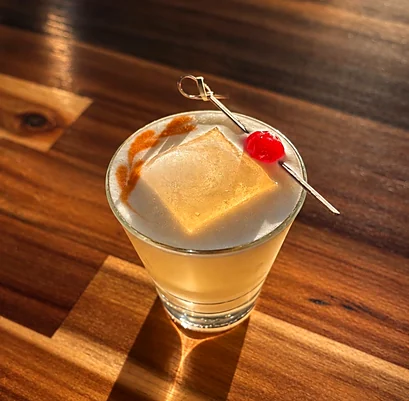Whiskey sour with a large ice cube and a cherry on a wooden bar top